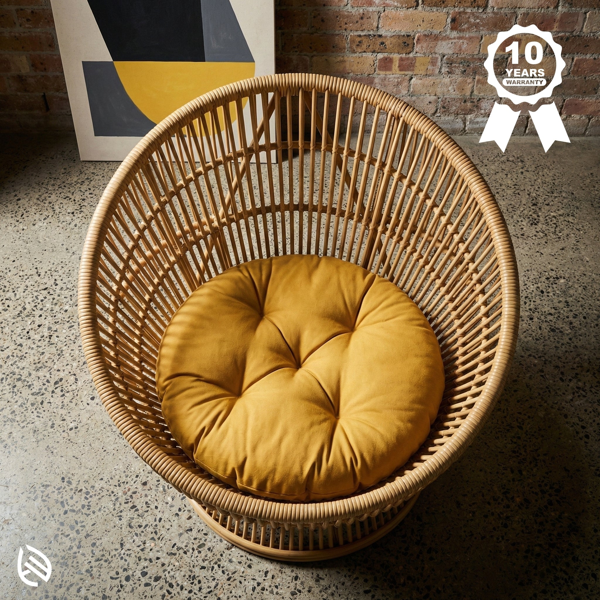 Wicker chair with a mustard yellow cushion on a textured floor, with a brick wall and abstract art in the background.